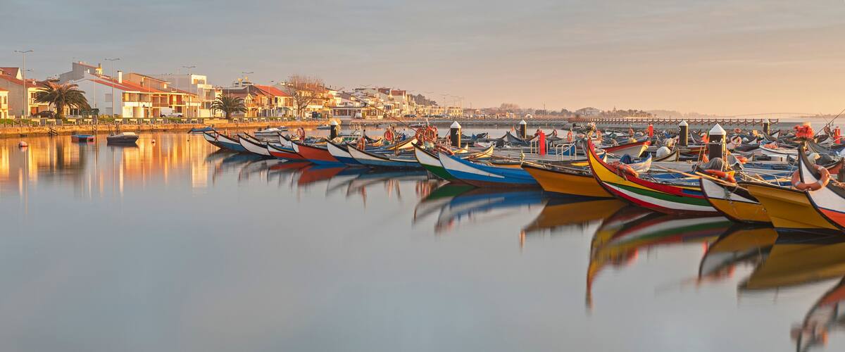 A calm harbor with colorful boats docked, reflecting in the water, and buildings in the background.