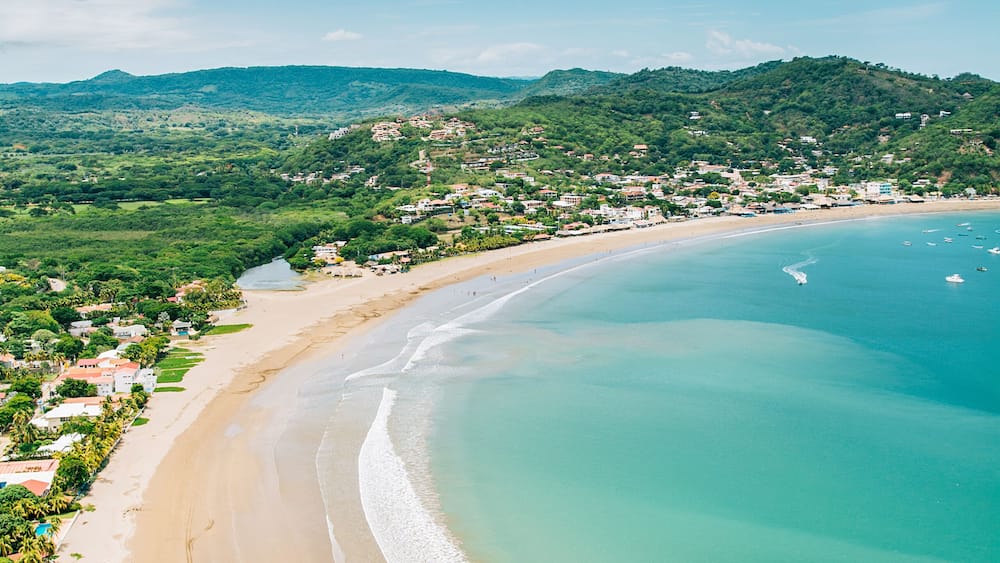 Eine Küstenlandschaft mit Sandstrand, klarem türkisblauem Wasser und sanften Hügeln mit verstreuten Gebäuden im Hintergrund.