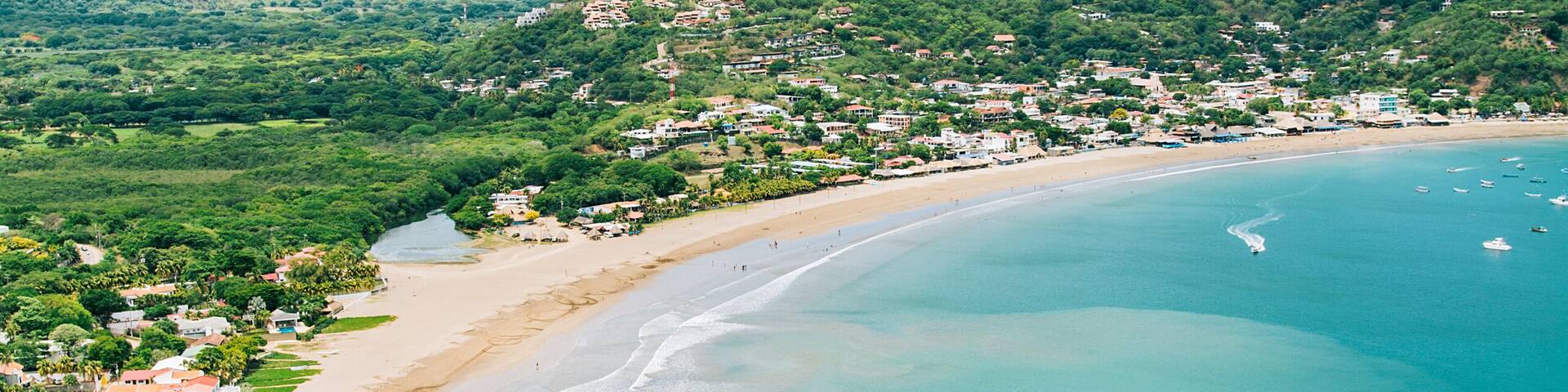 A coastal landscape with a sandy beach, clear turquoise waters, and a backdrop of hills with scattered buildings.