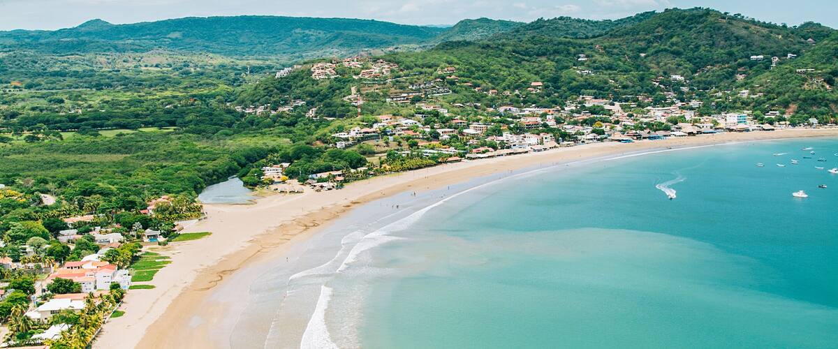 A coastal landscape with a sandy beach, clear turquoise waters, and a backdrop of hills with scattered buildings.