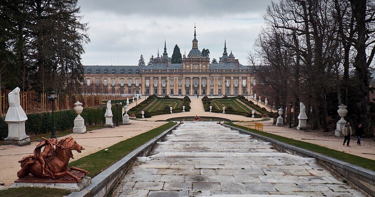 A grand palace with symmetrical gardens, a central statue, and a formal pathway leading to the entrance.