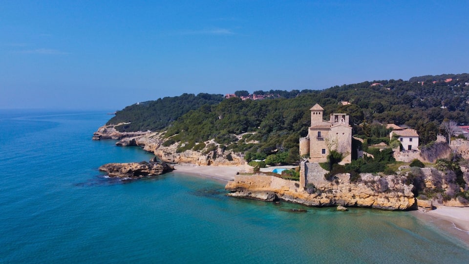 A coastal town with a historic stone building, clear turquoise waters, and a hilly backdrop.