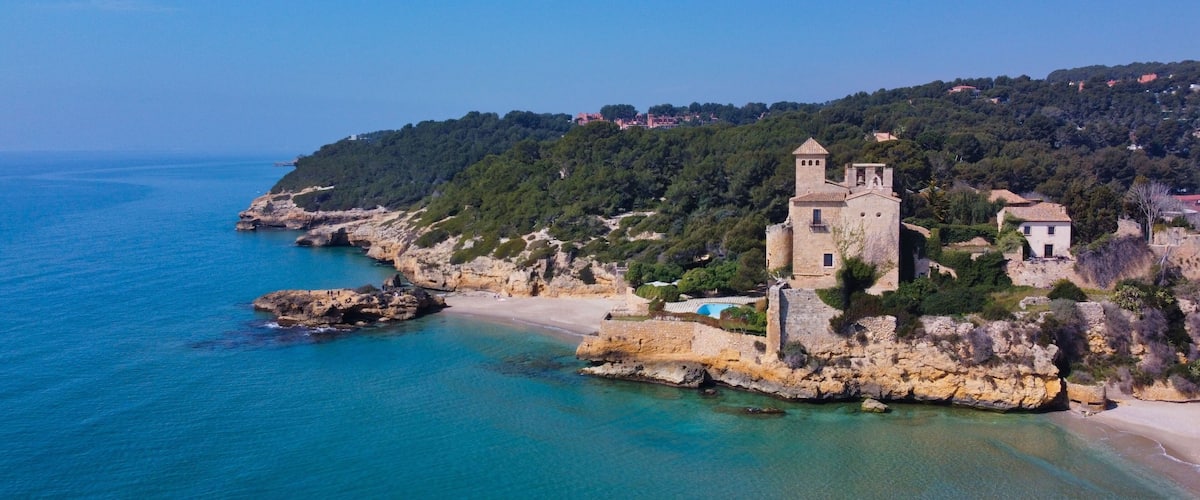 A coastal town with a historic stone building, clear turquoise waters, and a hilly backdrop.