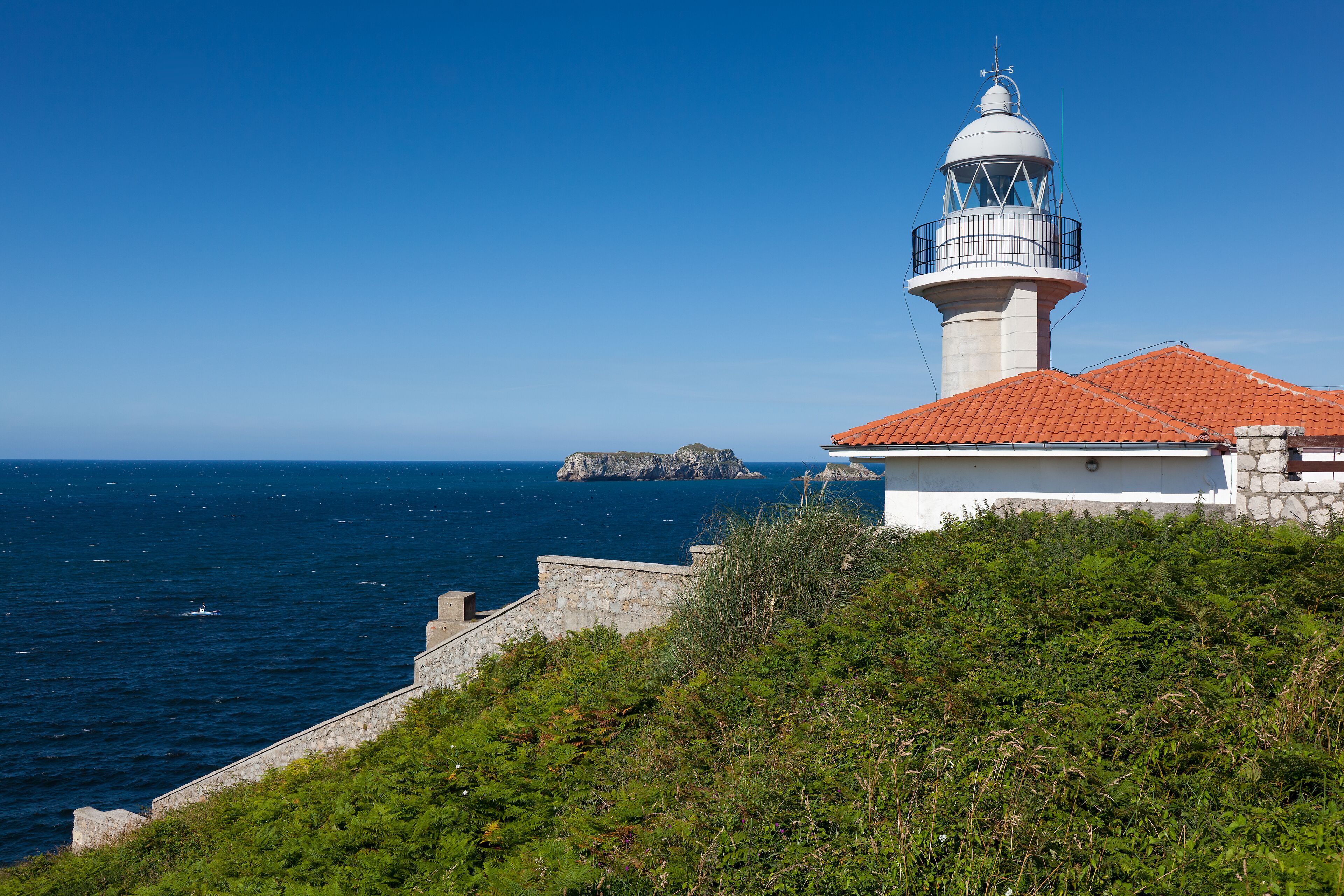 A lighthouse with a red roof, a stone wall, and a rocky outcrop in the background.