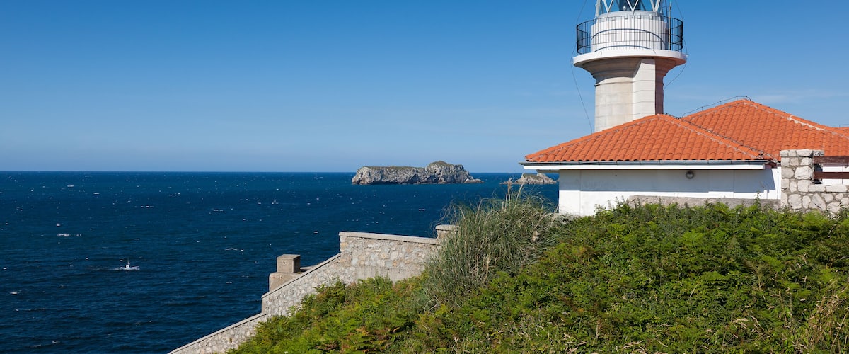 A lighthouse with a red roof, a stone wall, and a rocky outcrop in the background.