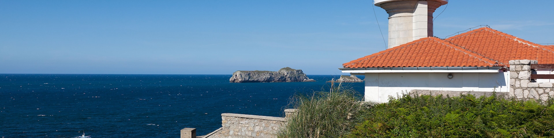 A lighthouse with a red roof, a stone wall, and a rocky outcrop in the background.