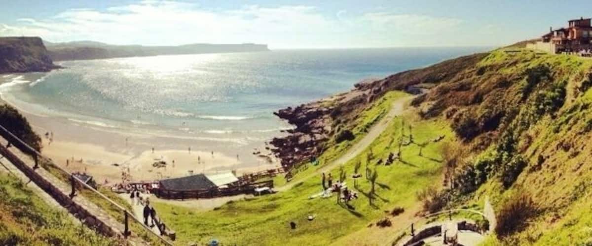 A coastal landscape with a beach, grassy hills, and a building on a hill.
