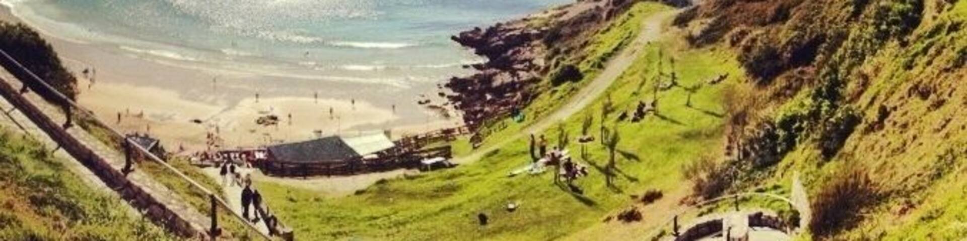 A coastal landscape with a beach, grassy hills, and a building on a hill.