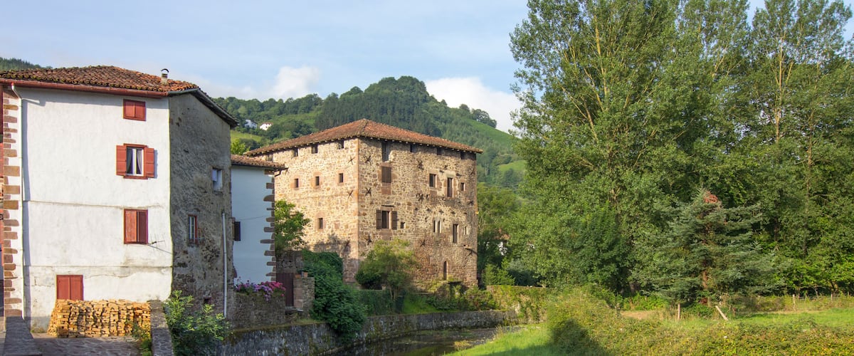 A stone building with a tiled roof, a river, and lush greenery.