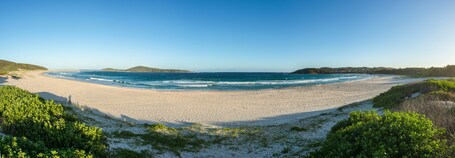 A wide sandy beach with gentle waves, surrounded by green hills and vegetation.