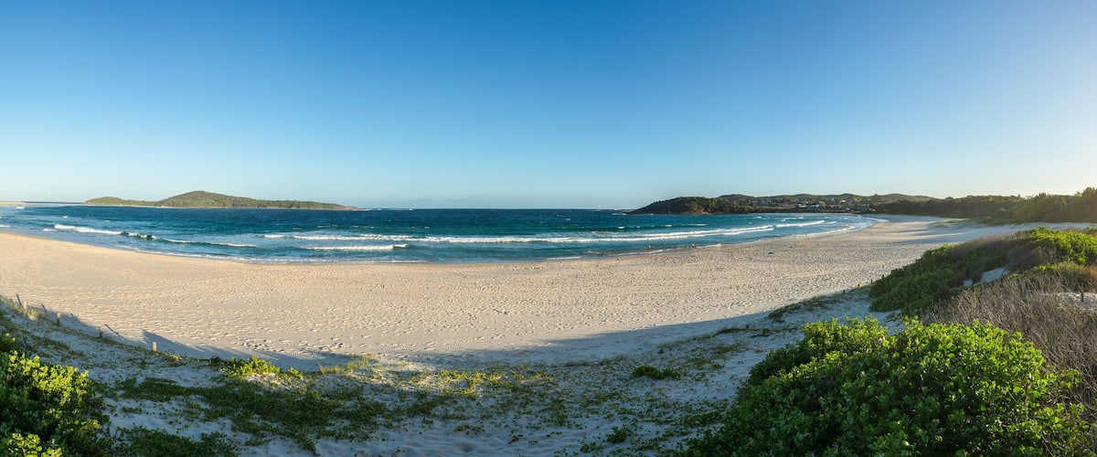 A wide sandy beach with gentle waves, surrounded by green hills and vegetation.