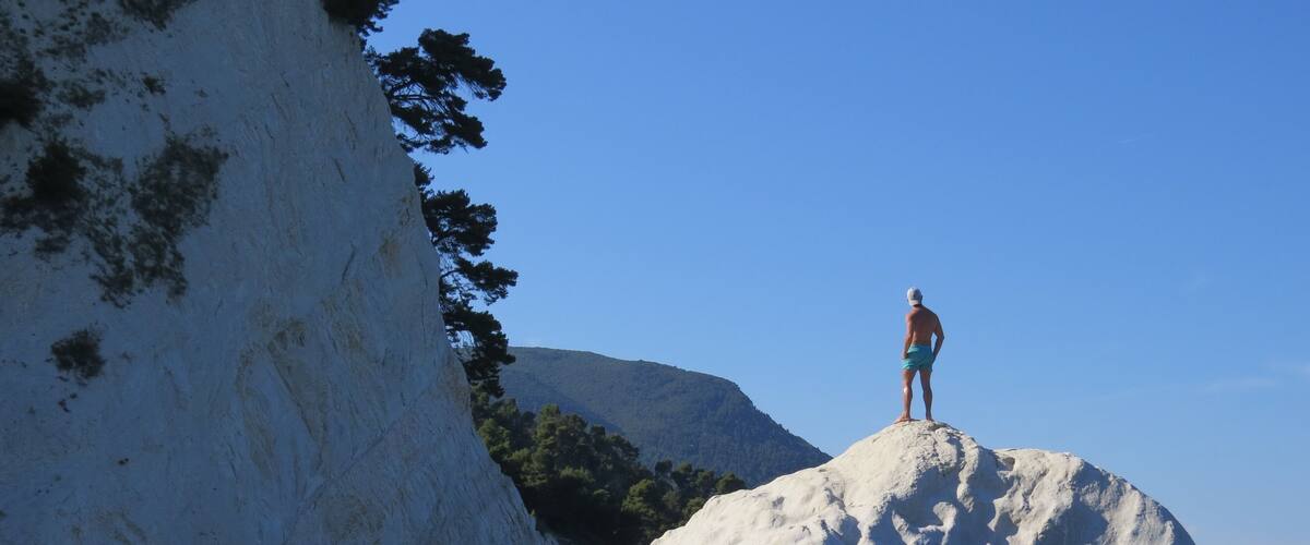 A person standing on a large white rock in clear water, with a cliff and trees in the background.
