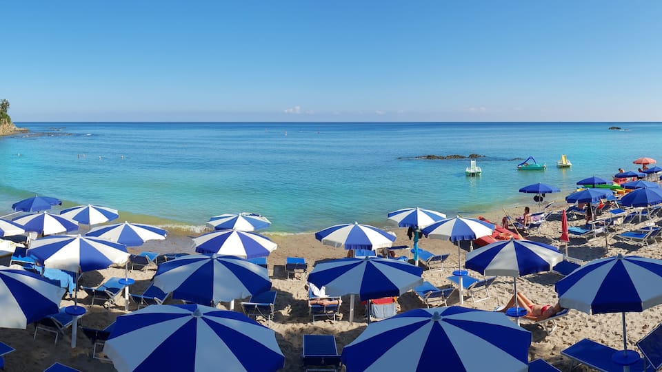 A beach with numerous blue and white striped umbrellas, clear turquoise water, and a rocky shoreline.