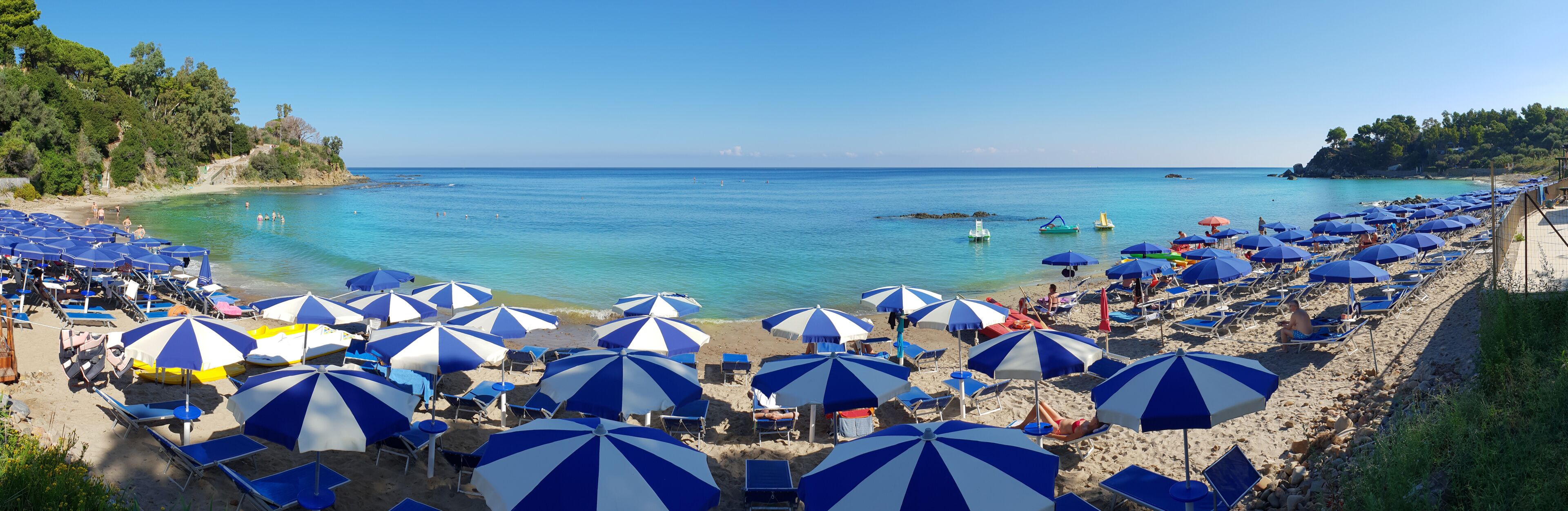 A beach with numerous blue and white striped umbrellas, clear turquoise water, and a rocky shoreline.