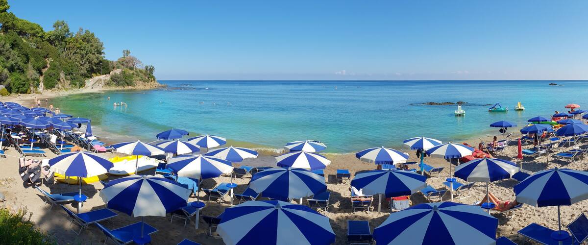 A beach with numerous blue and white striped umbrellas, clear turquoise water, and a rocky shoreline.