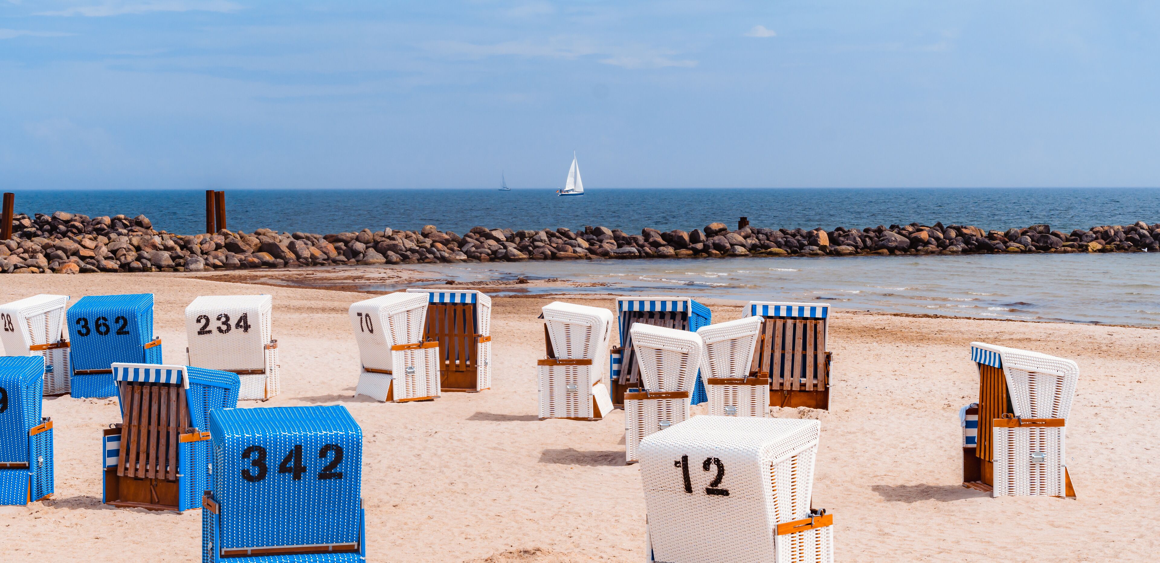 Beach chairs with numbers on a sandy beach, a rocky breakwater, and a sailboat in the distance.