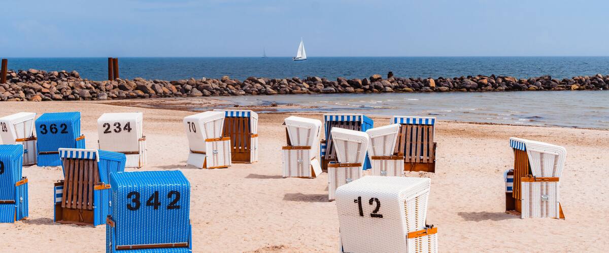 Beach chairs with numbers on a sandy beach, a rocky breakwater, and a sailboat in the distance.