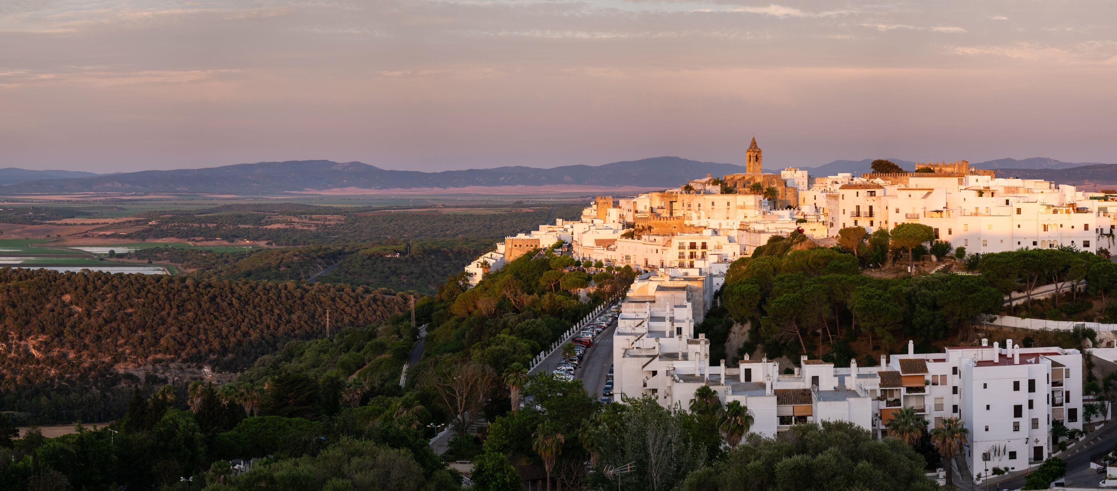 A coastal town with white buildings, a prominent church tower, and a hilly backdrop.
