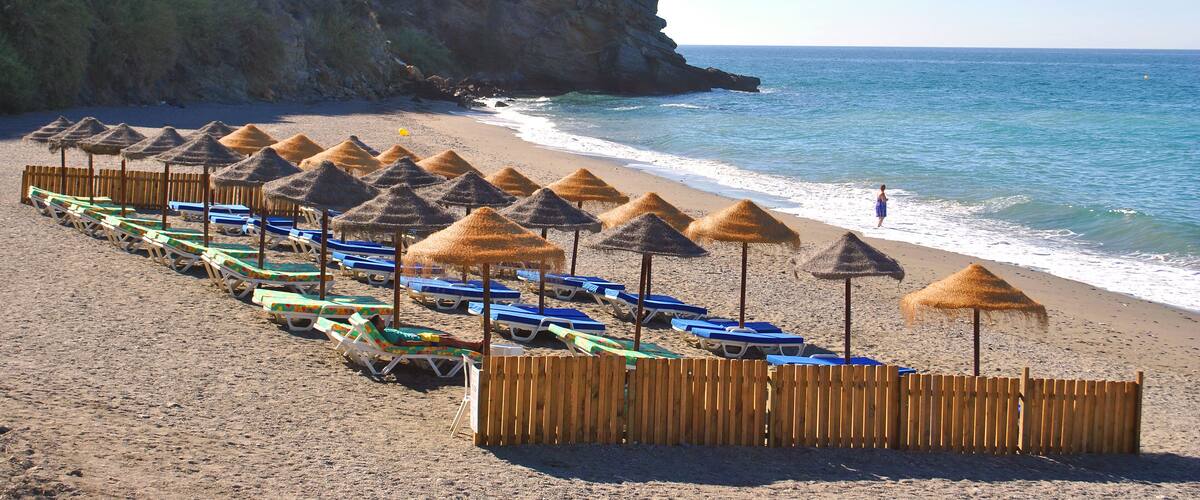 A beach with rows of sun umbrellas and lounge chairs, a rocky cliff, and the ocean.