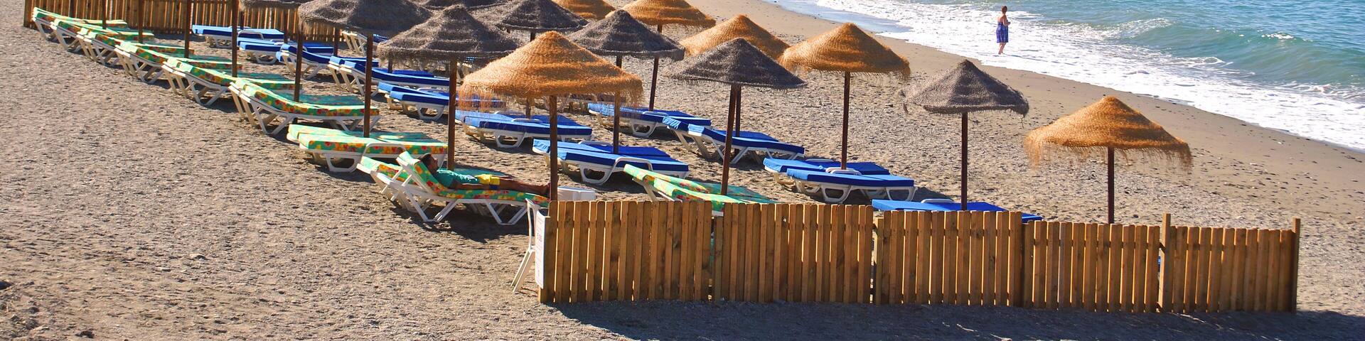 A beach with rows of sun umbrellas and lounge chairs, a rocky cliff, and the ocean.