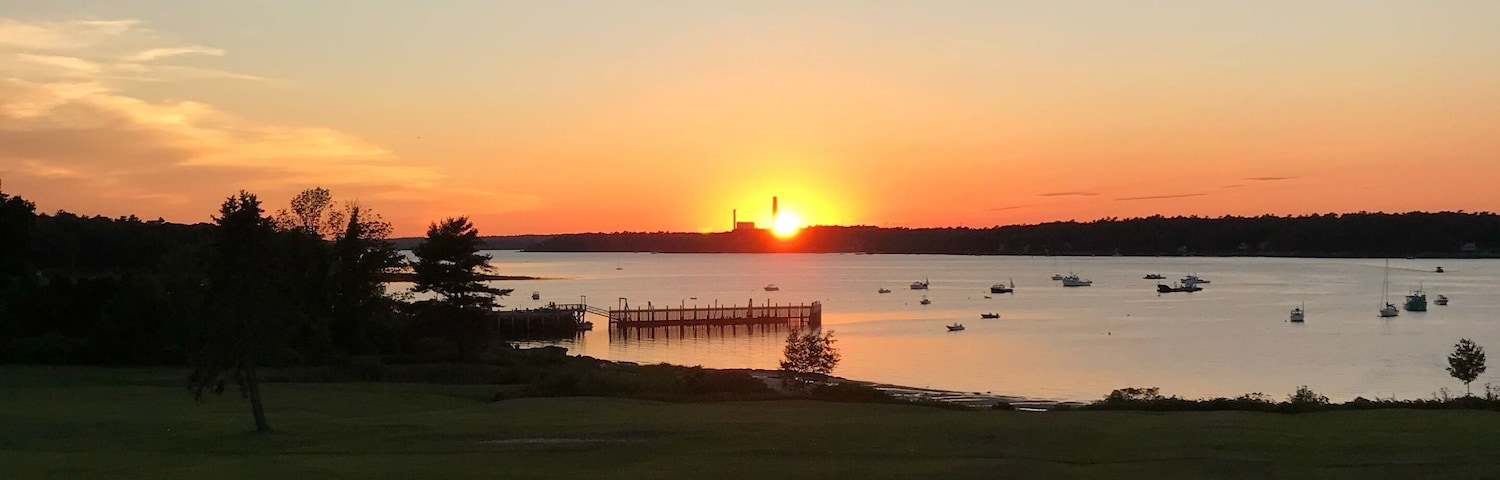 Um pôr do sol sobre um lago calmo, com barcos, um cais e um campo gramado.