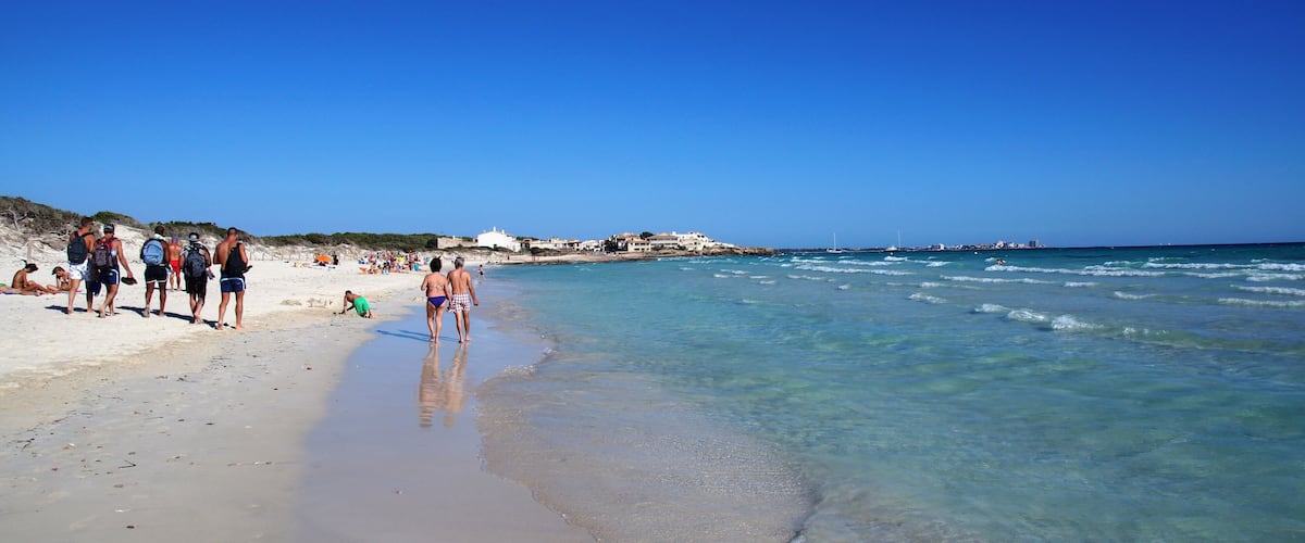 Una playa con agua azul cristalina, orilla arenosa y algunas personas caminando por la orilla.