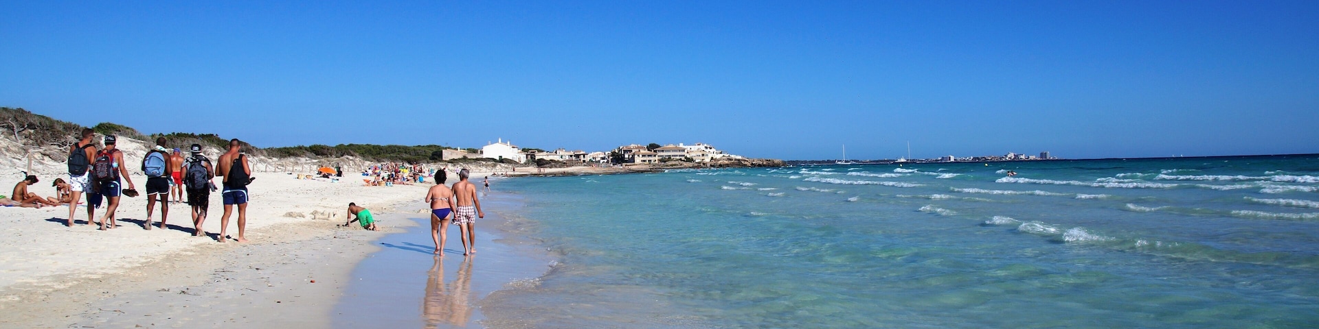 Una playa con agua azul cristalina, orilla arenosa y algunas personas caminando por la orilla.