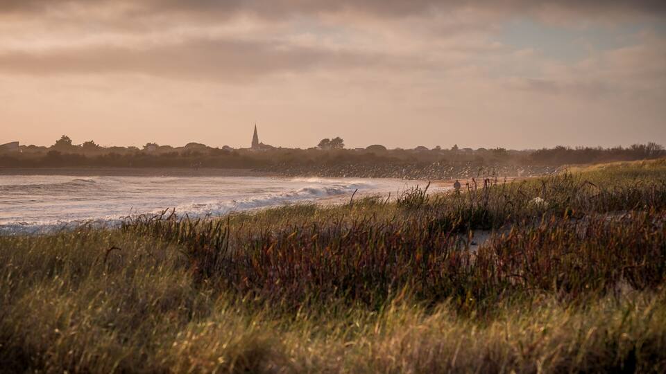 A coastal landscape with waves, grassy dunes, and a distant church spire.