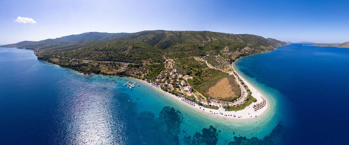 An aerial view of a coastal island with a beach, clear blue waters, and lush green hills.