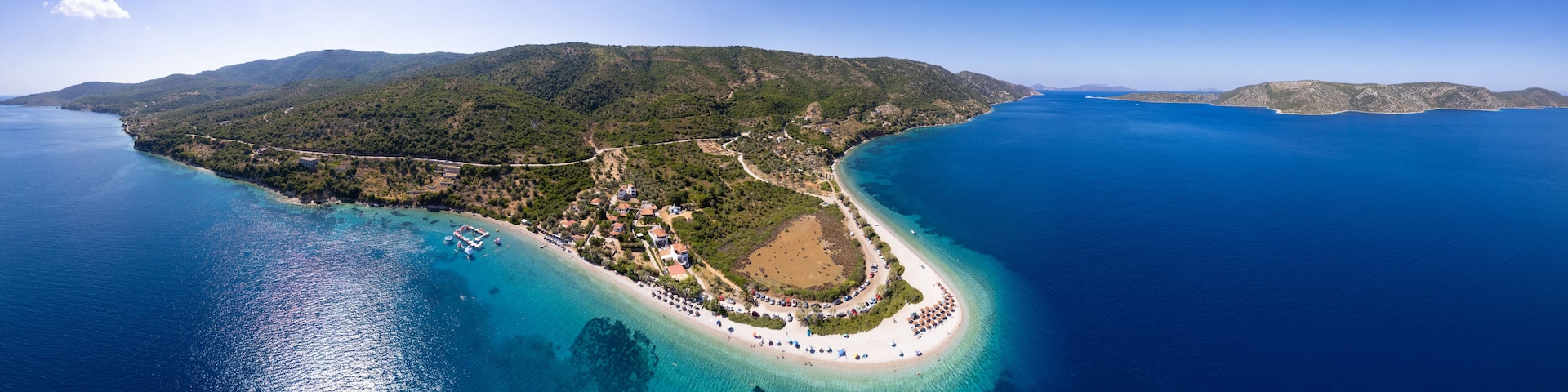 An aerial view of a coastal island with a beach, clear blue waters, and lush green hills.