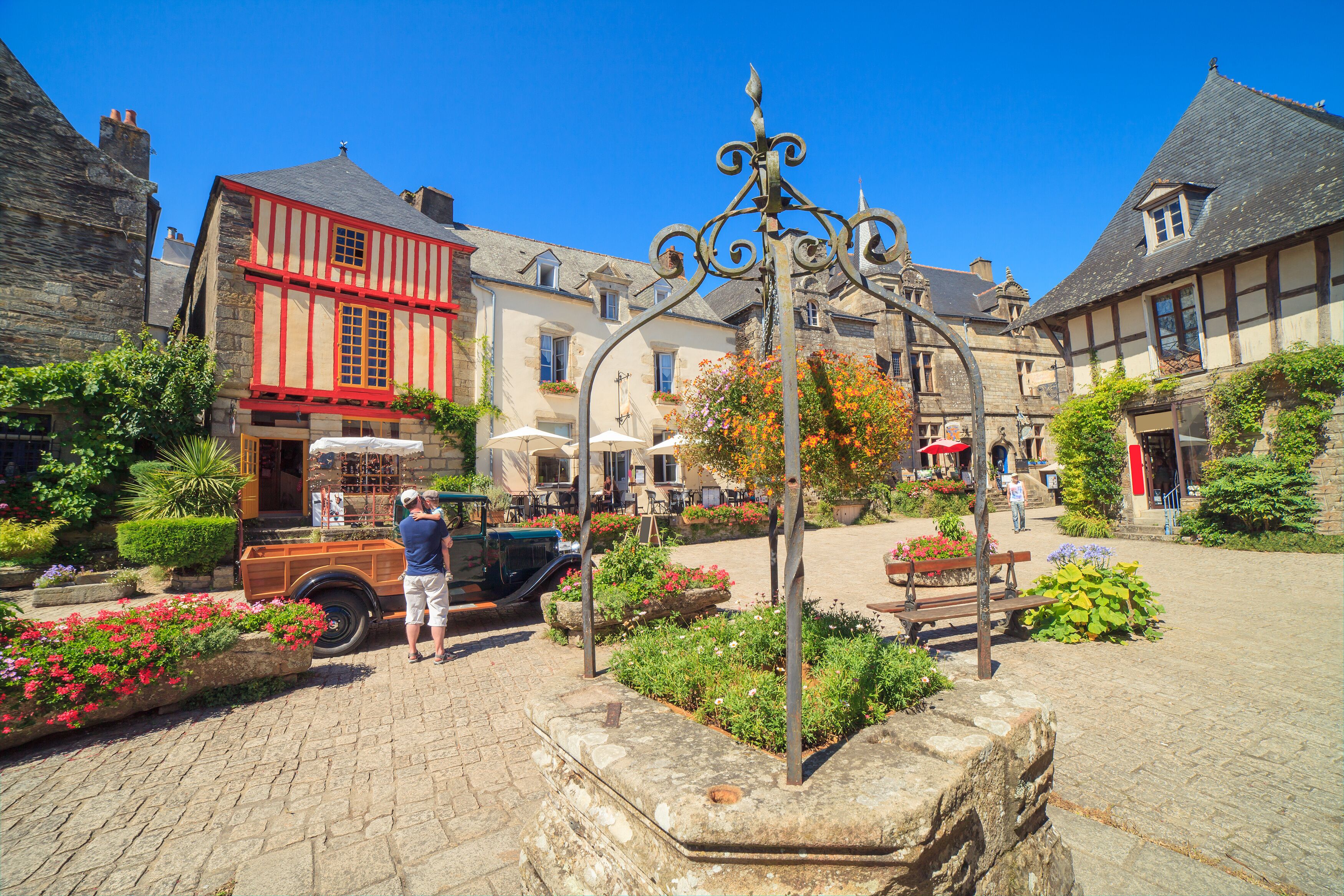 A cobblestone street with historic buildings, a vintage car, and a decorative ironwork structure.