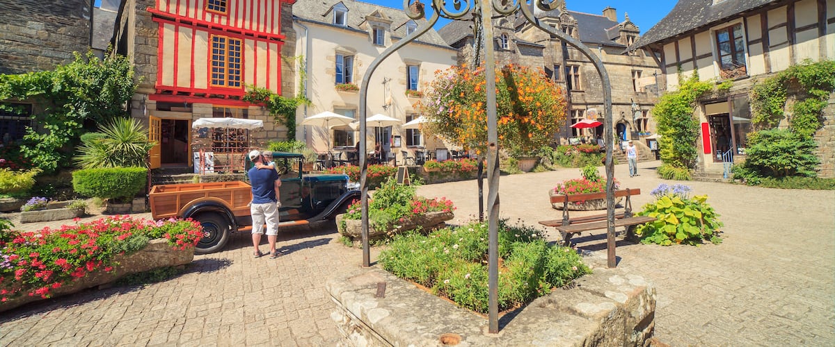 A cobblestone street with historic buildings, a vintage car, and a decorative ironwork structure.