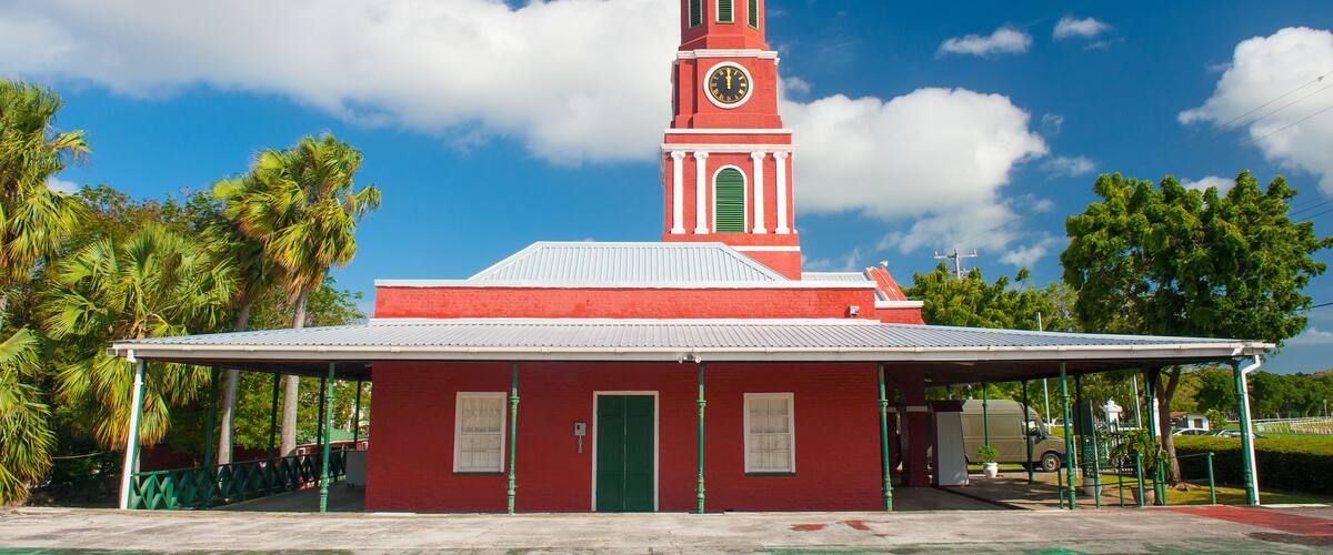 A red church with a clock tower and a green dome, surrounded by palm trees and a clear blue sky.