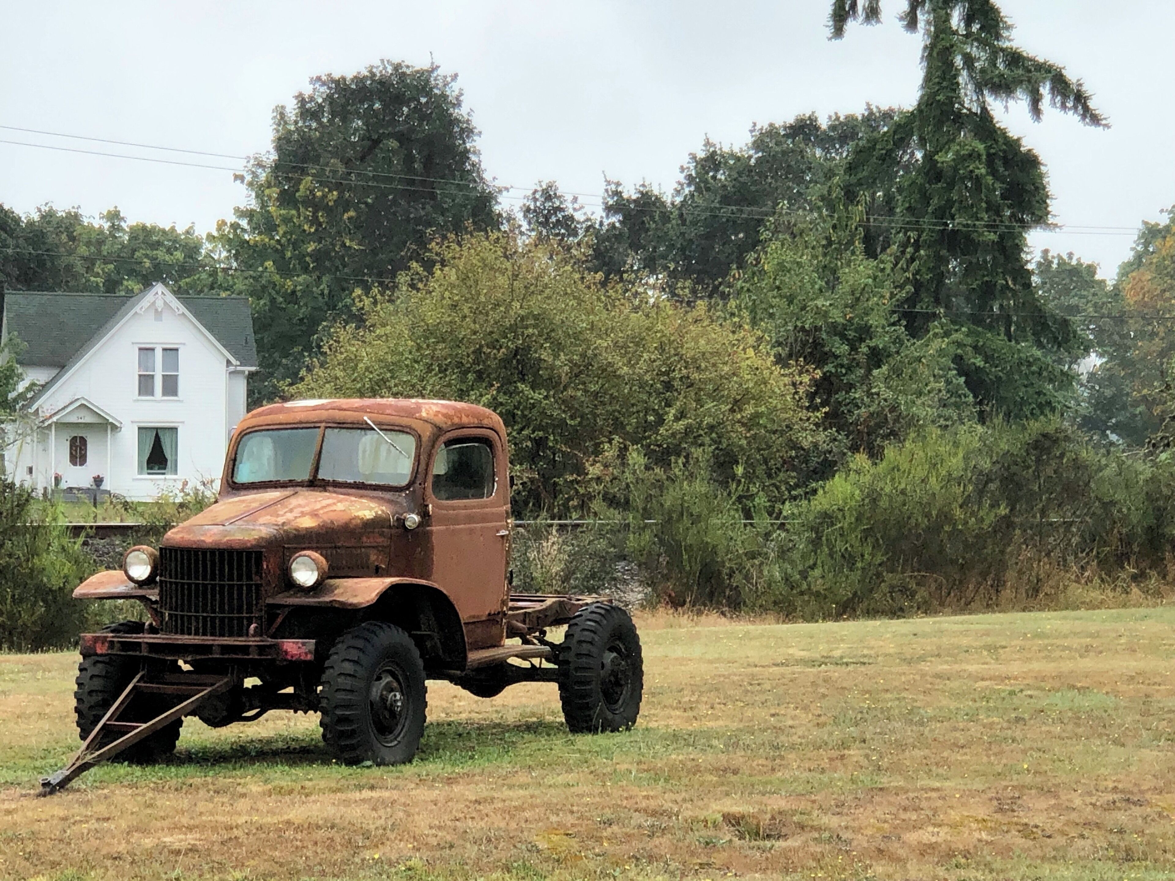 An old rusty truck parked in a grassy field with a white house and dense trees in the background.