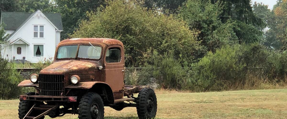 An old rusty truck parked in a grassy field with a white house and dense trees in the background.