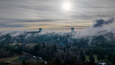 Aerial view of twin cooling towers emerge through the dense fog, piercing the skyline above verdant forest, Elma, Washington, United States.