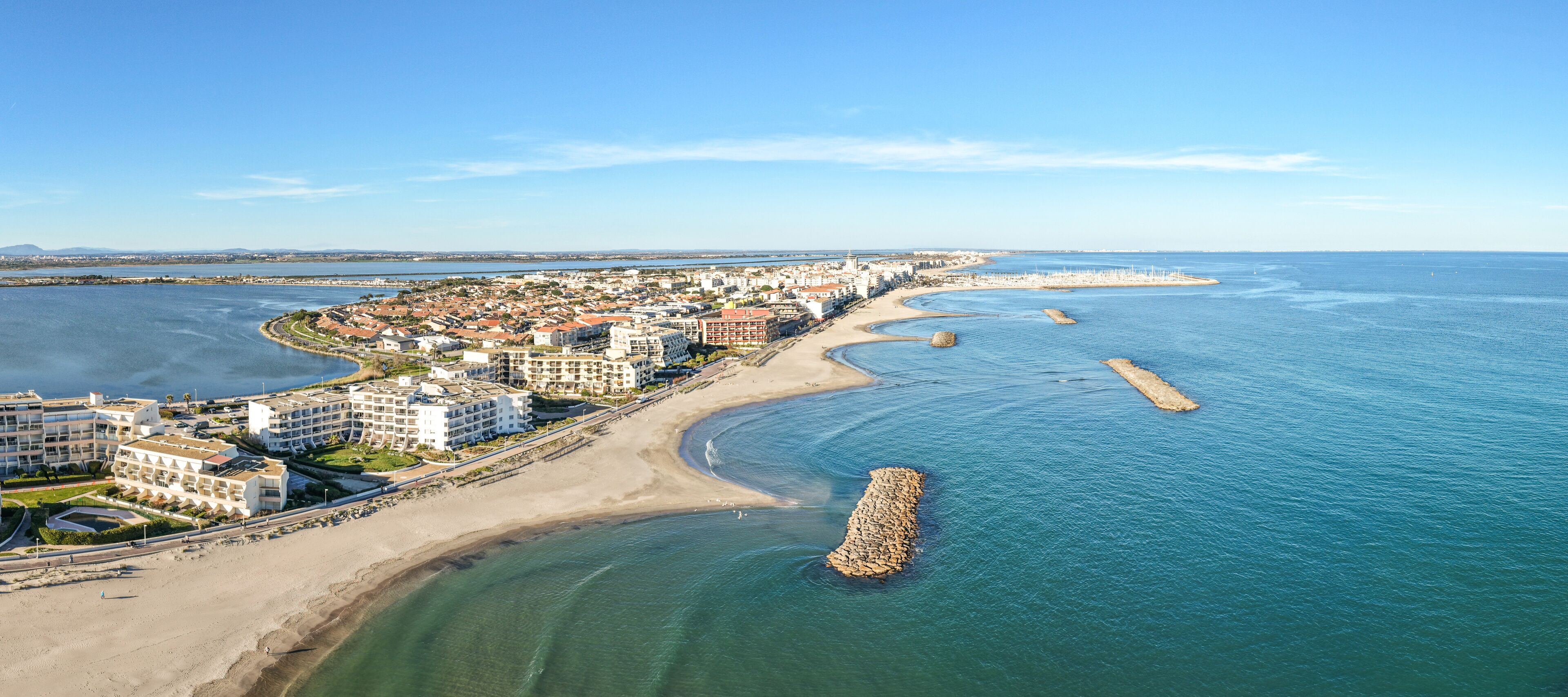 A coastal cityscape with a sandy beach, a marina, and a prominent breakwater.
