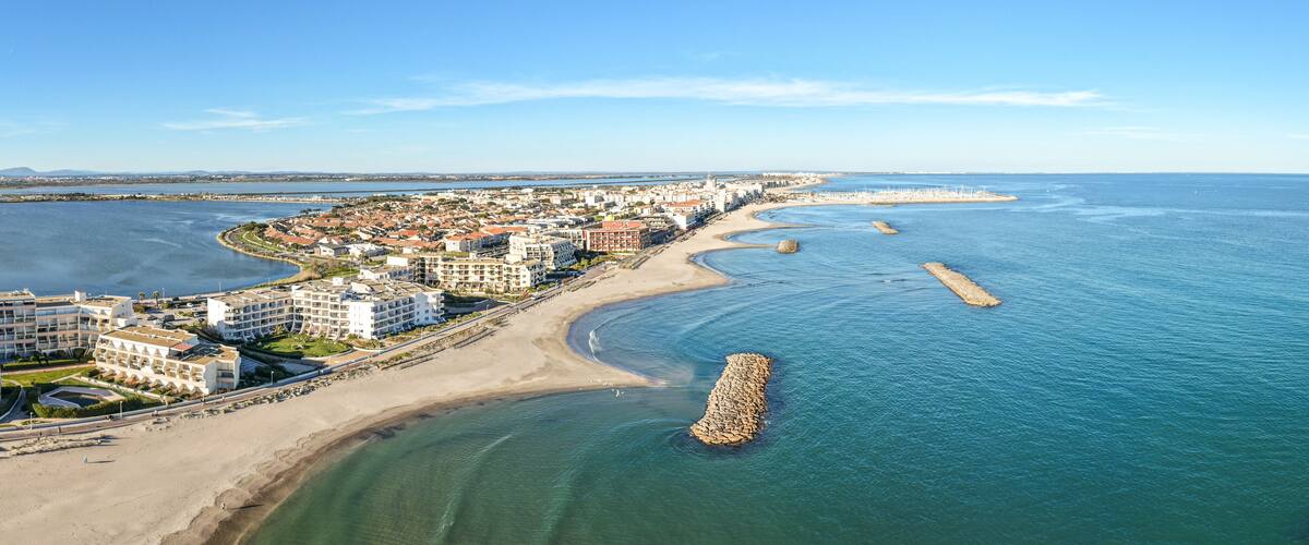 A coastal cityscape with a sandy beach, a marina, and a prominent breakwater.