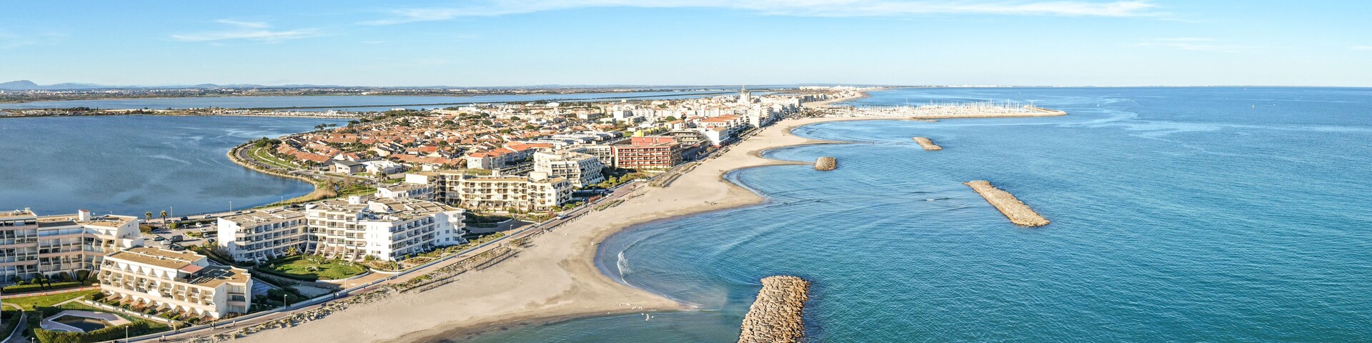 A coastal cityscape with a sandy beach, a marina, and a prominent breakwater.