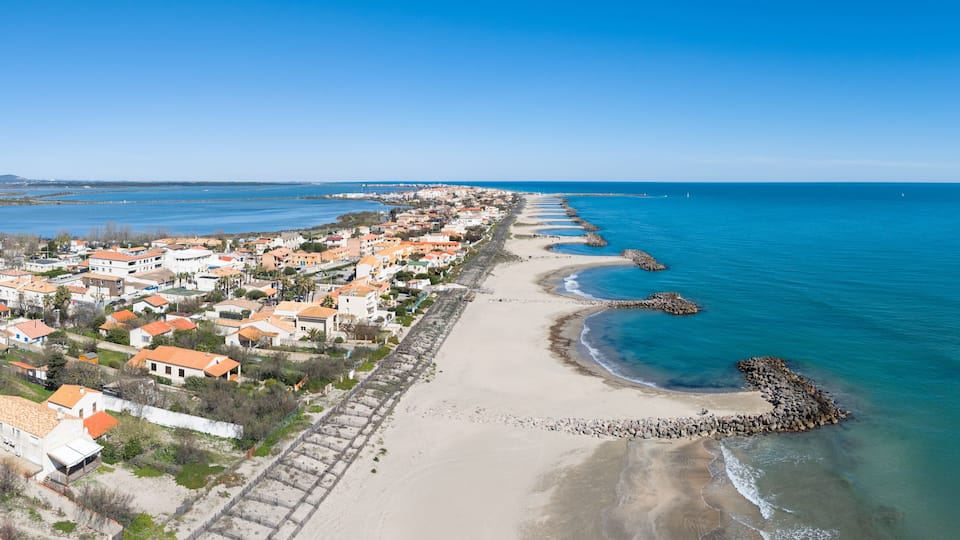 A coastal town with a long sandy beach, a rocky breakwater, and residential buildings with orange roofs.
