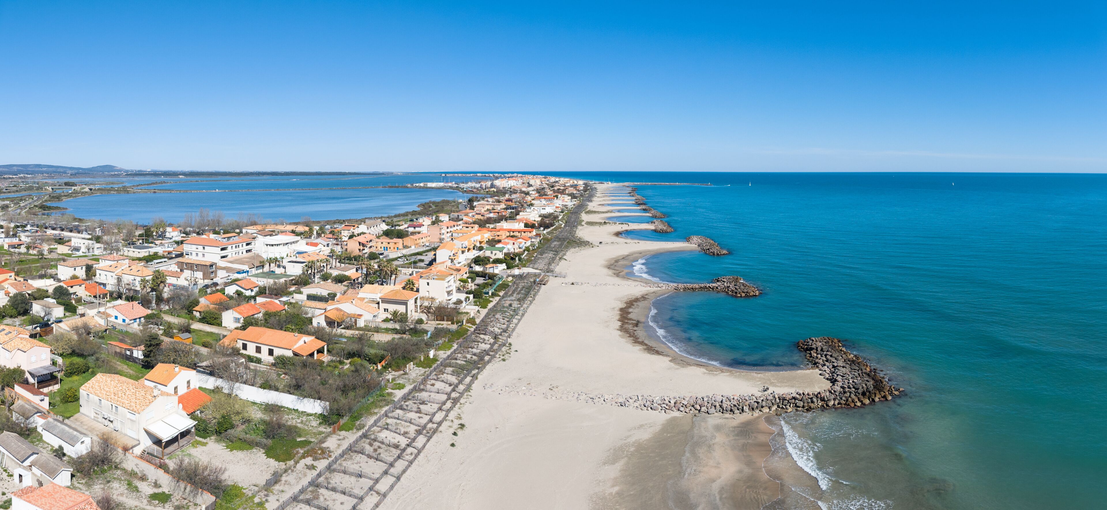 A coastal town with a long sandy beach, a rocky breakwater, and residential buildings with orange roofs.