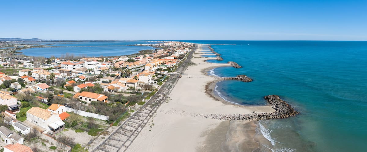 A coastal town with a long sandy beach, a rocky breakwater, and residential buildings with orange roofs.