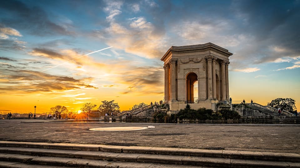 A grand arch monument with steps leading up to it, set against a sunset sky.