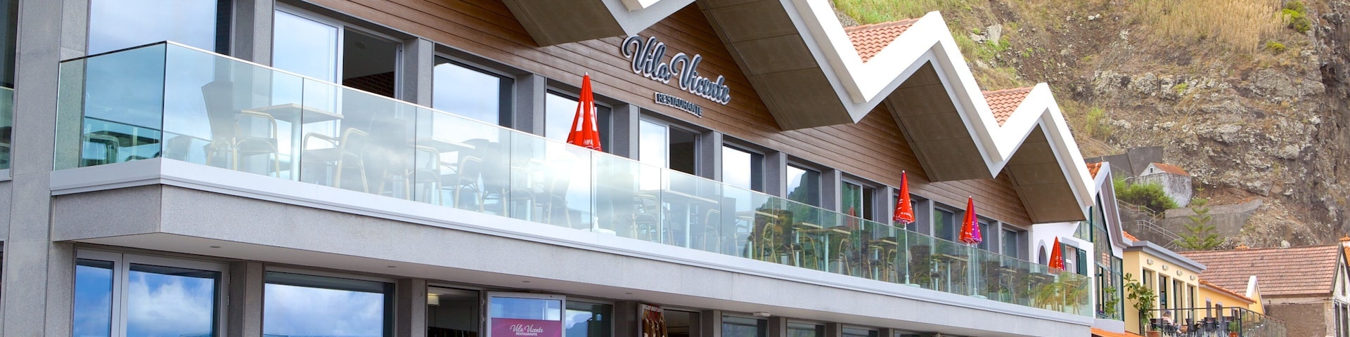 A coastal restaurant with outdoor seating, red umbrellas, and a cliffside view.