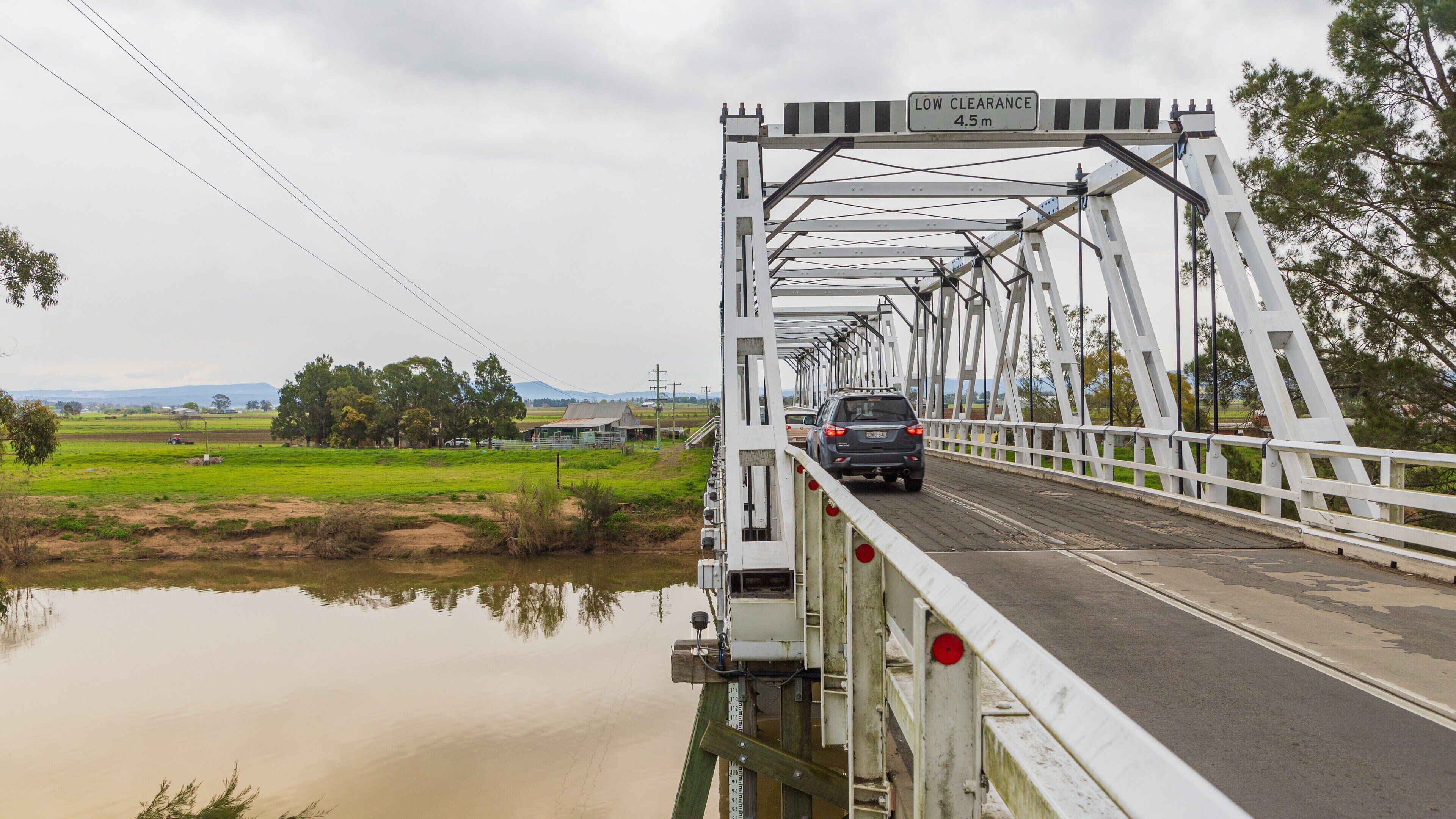 A bridge with a low clearance sign of 4.5 meters, a car crossing, and a rural landscape with fields and a body of water.