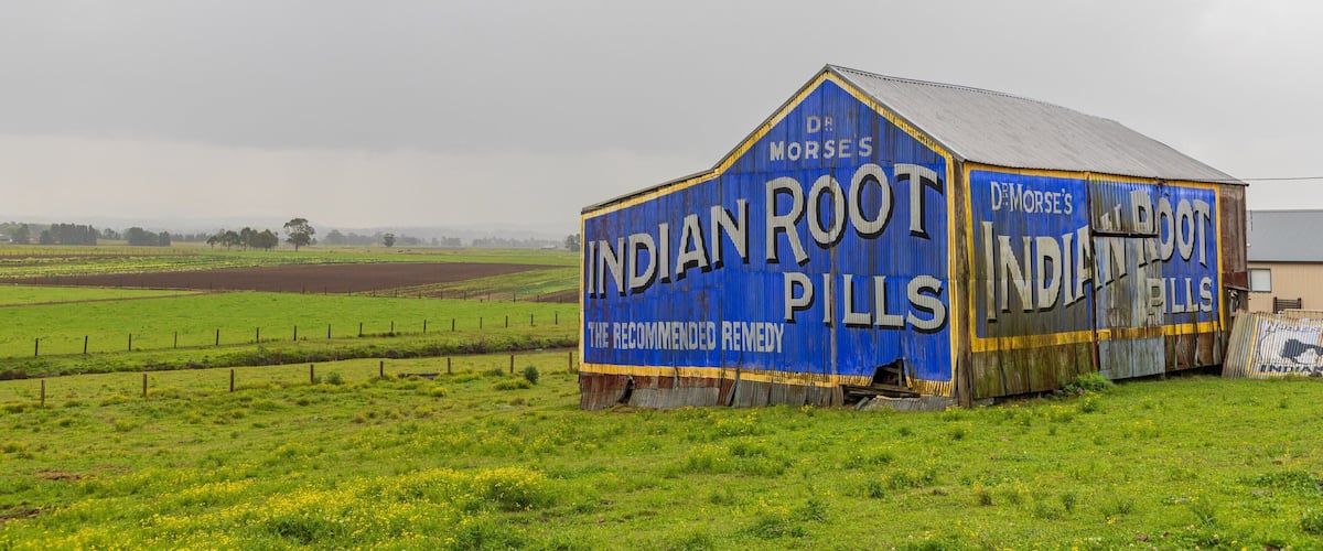 A dilapidated barn with 'Indian Root Pills' written on its side, surrounded by a green field.