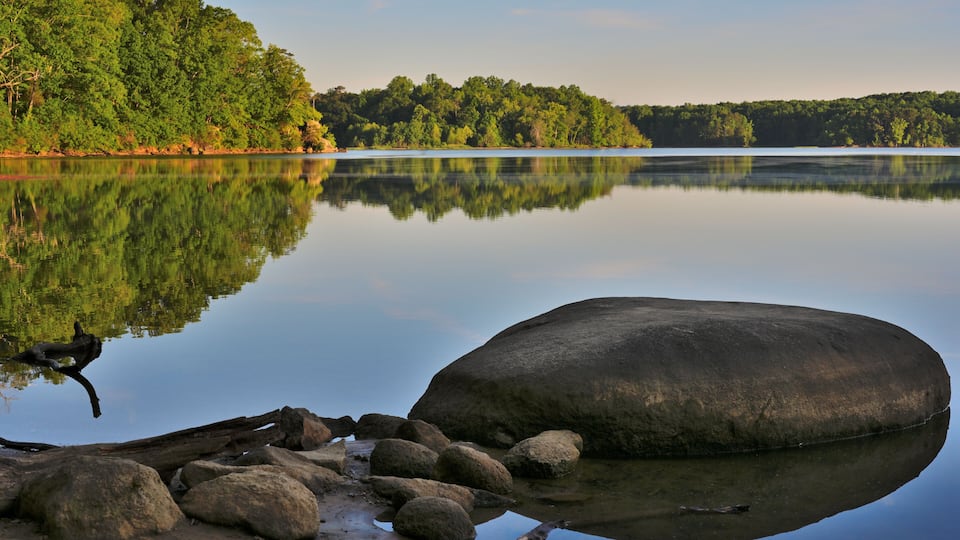 A calm lake with a large rock in the foreground and a forested shoreline in the background.