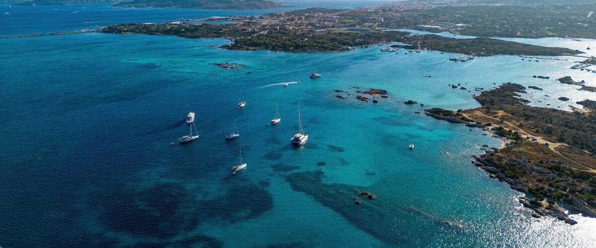 An aerial view of a coastal area with clear turquoise waters, several sailboats, and a distant cityscape.