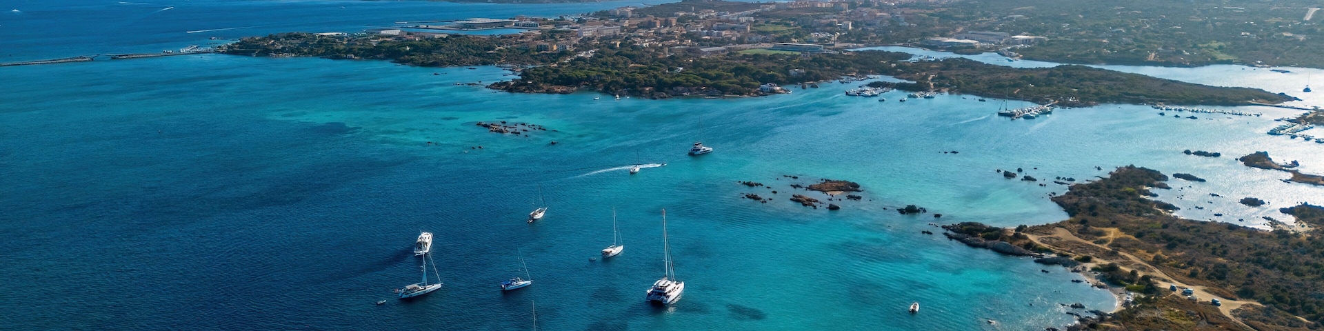 An aerial view of a coastal area with clear turquoise waters, several sailboats, and a distant cityscape.