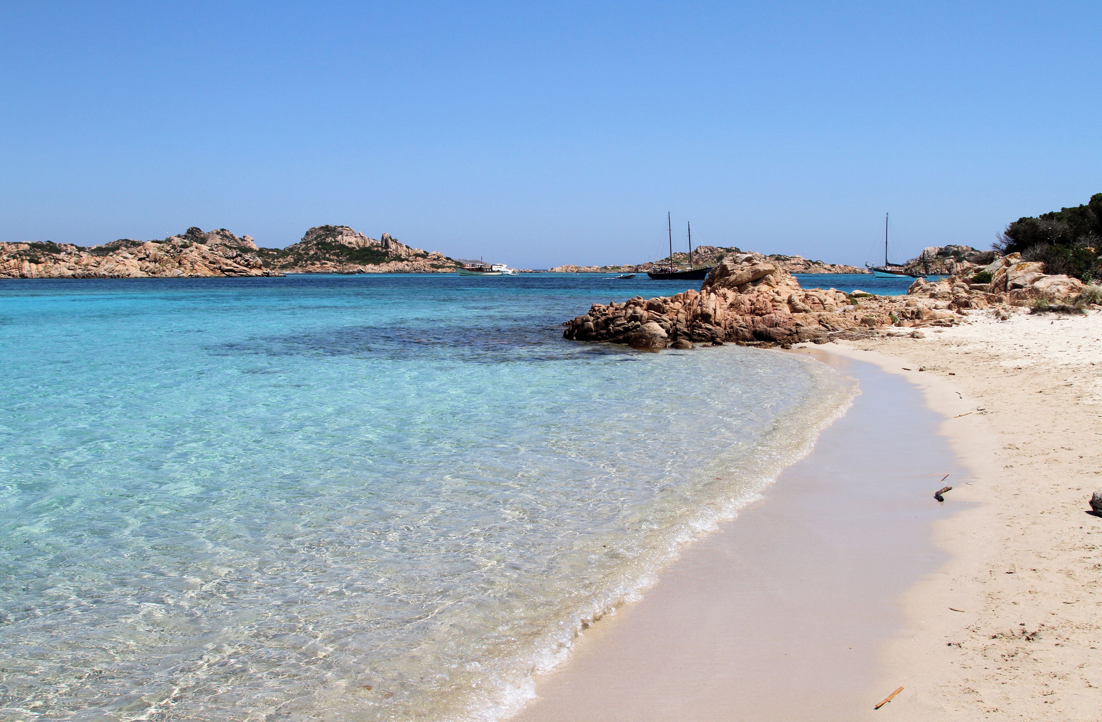 A clear turquoise sea, sandy beach, rocky outcrops, and distant sailboats.